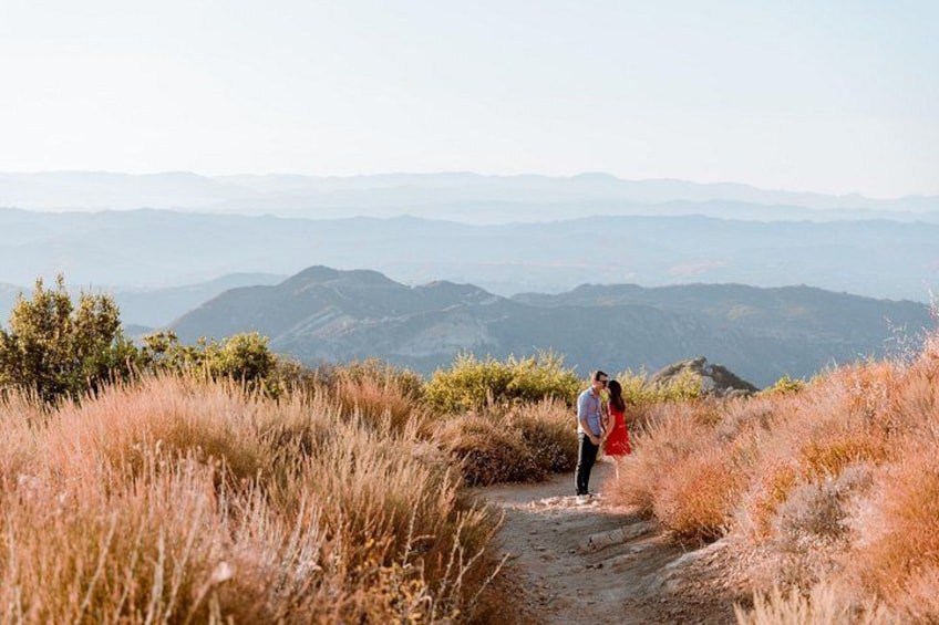 A couple walking on a dirt path surrounded by dry grass and hills in the distance. 4