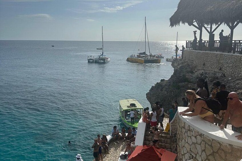 A coastal scene with a rocky shore, a thatched-roof structure, and boats on the water. 3