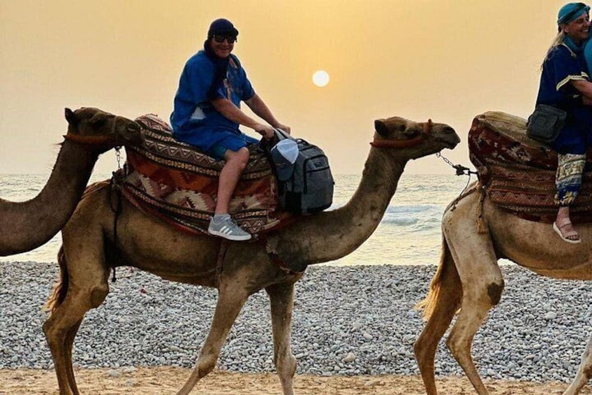 Two camels with riders on a pebbled beach at sunset. 5