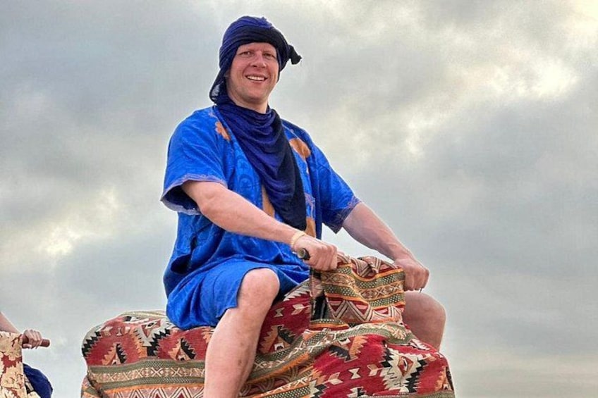 Een persoon die op een kameel rijdt, met een versierd zadel, gekleed in een blauwe gewaad en hoofdbedekking, voor een bewolkte lucht. 2