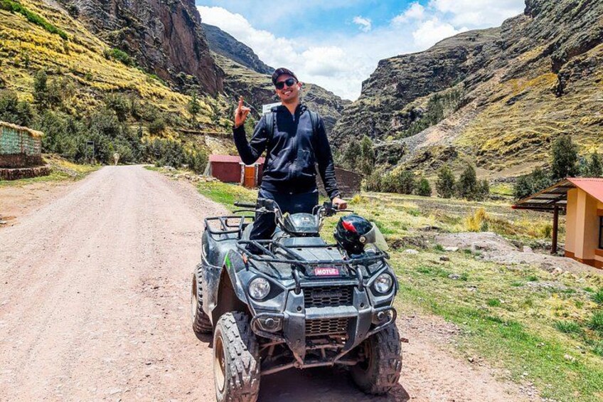 Une personne conduit un quad sur un chemin de terre, dans un paysage rural avec des montagnes et un petit bâtiment en arrière-plan. 2