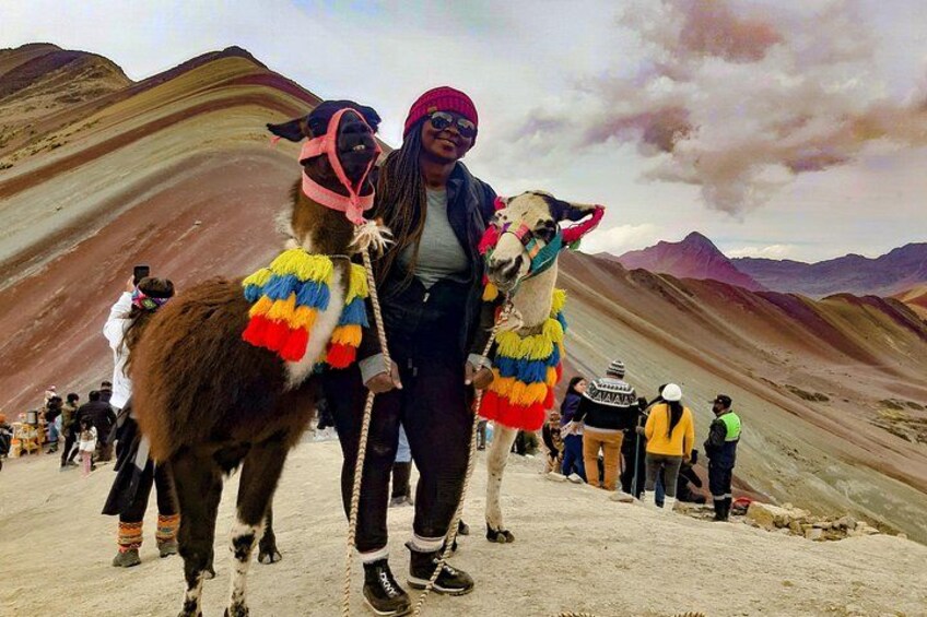 Deux lamas portant des couvertures colorées, une personne au premier plan et un paysage montagneux avec des personnes à l’arrière-plan. 5