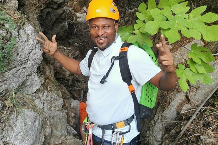 A person wearing a helmet and backpack, standing next to a rocky terrain with green foliage. 2