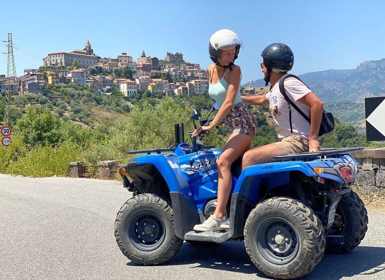 Two people on a blue ATV on a road with a view of a town and mountains in the background. 1