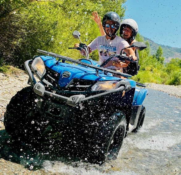 Two people on a blue ATV driving through a shallow stream, splashing water. 4