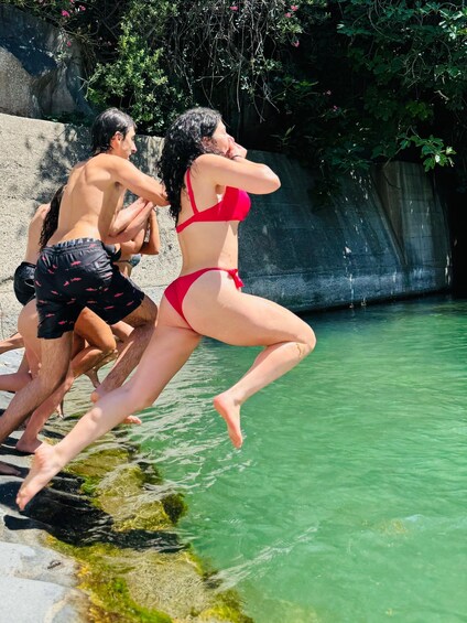 People jumping into a clear green pool surrounded by rocks and greenery. 3