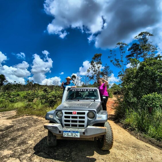 A silver SUV on a dirt road with two people standing on the back, surrounded by greenery and a clear blue sky. 2