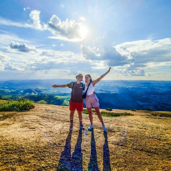 Two people standing on a rocky outcrop with their arms outstretched, overlooking a vast landscape with a clear sky and distant cityscape. 4