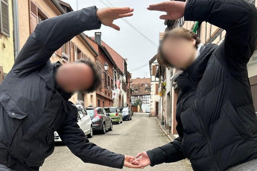Two people in black jackets making a hand gesture in a narrow street with parked cars and buildings. 2