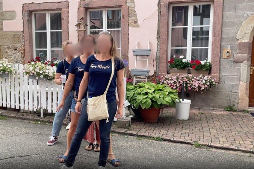 Drei Personen gehen auf einer Straße entlang, ein weißes Zaunfeld und ein Gebäude mit Blumenkästen. 4