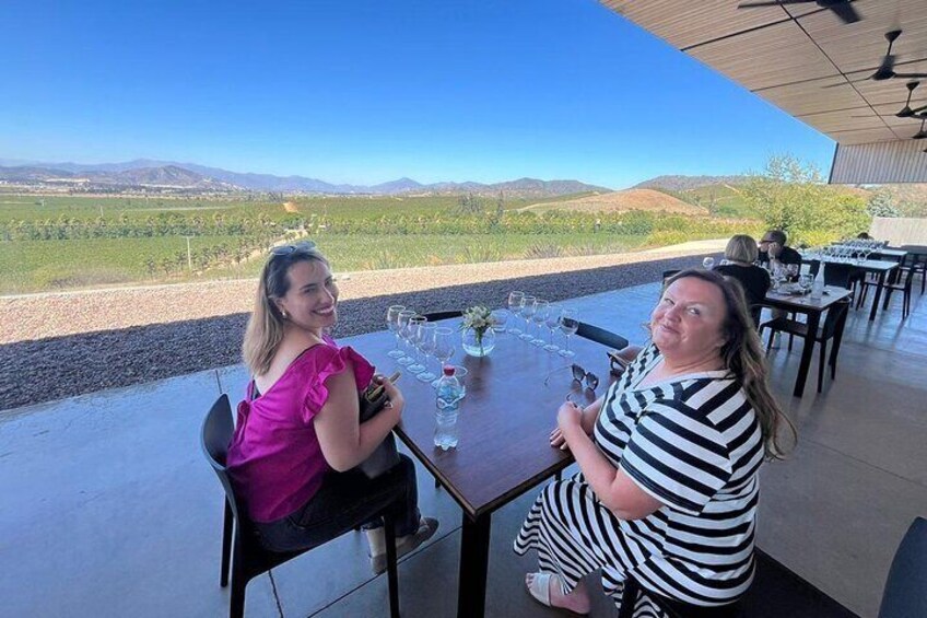 Two women sitting at an outdoor table with a view of a vineyard and hills in the background. 5