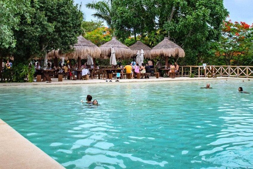 A swimming pool with people swimming and sunbathing, surrounded by thatched umbrellas and trees. 4