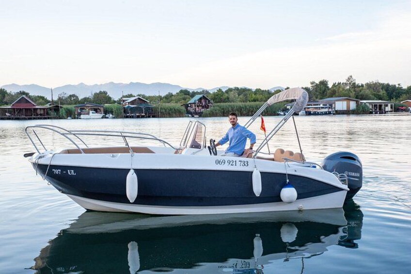 A motorboat with a cabin and outboard motor, docked on calm water with houses and mountains in the background. 1