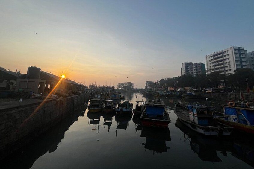 A calm river with boats docked along the shore, buildings in the background, and the sun setting on the horizon. 5