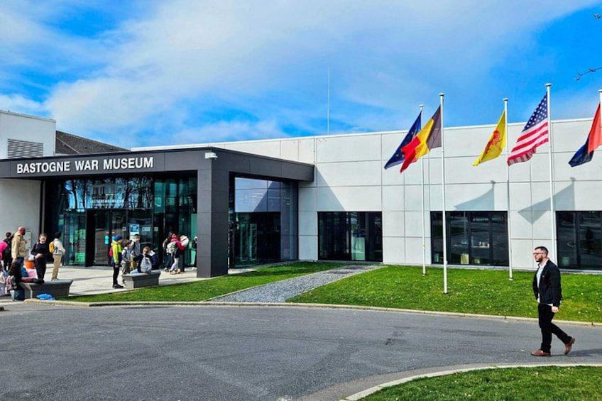 Das Bastogne War Museum mit Flaggen, Spaziergängern und einem modernen Gebäude. 2