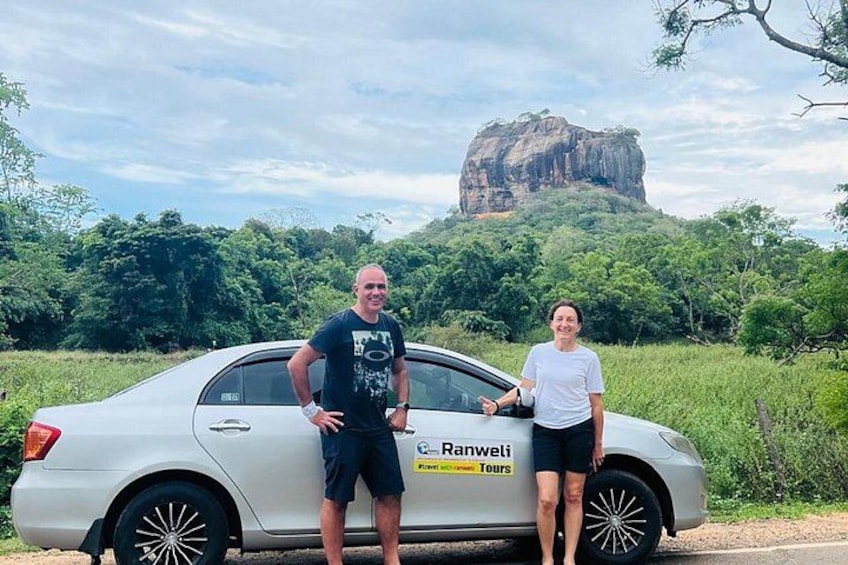 A white car with a license plate, two people standing beside it, and a large rock formation in the background. 1