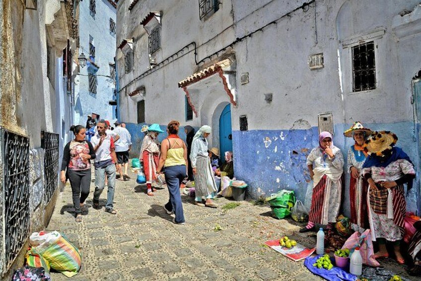 A narrow street with people walking, some carrying items, and buildings with blue and white facades. 2