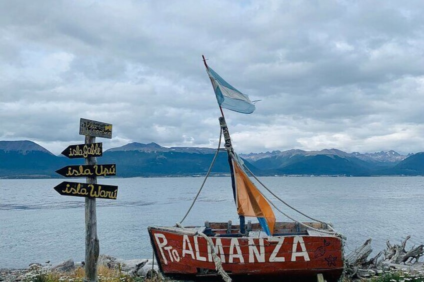 A boat named "P. ALMANZA" with a sail, a signpost with directions to islands, and a lake with mountains in the background. 2
