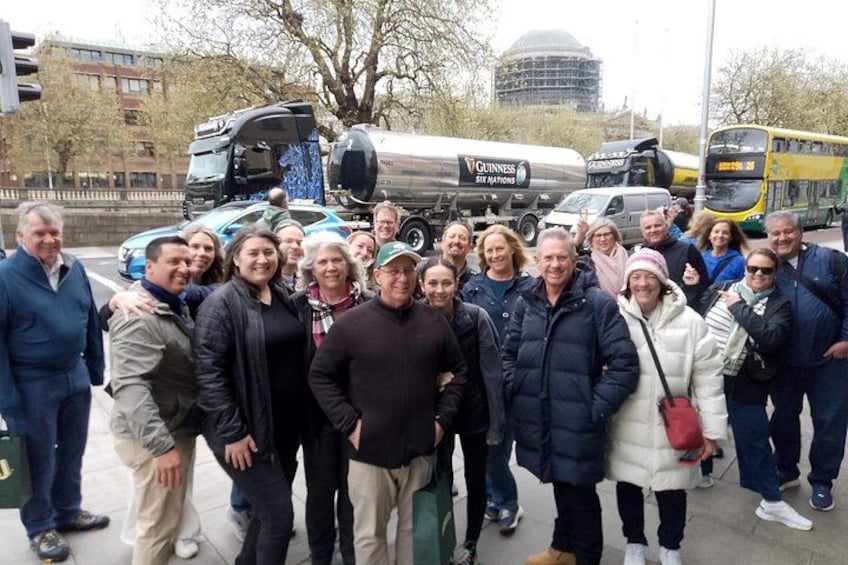A group of people standing on a sidewalk with a bus and a truck in the background. 1