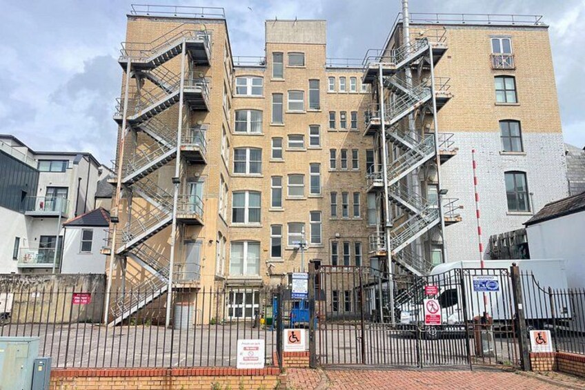 Un immeuble à plusieurs étages doté d’échelles de secours extérieures, d’une entrée sécurisée par un portail et d’une enseigne bleue. 4