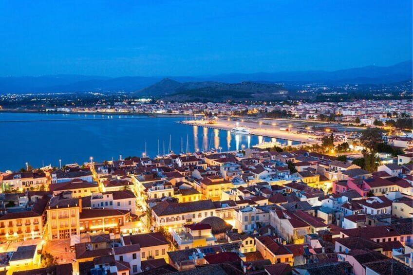 A coastal cityscape at dusk with illuminated buildings, a marina, and a bridge. 1