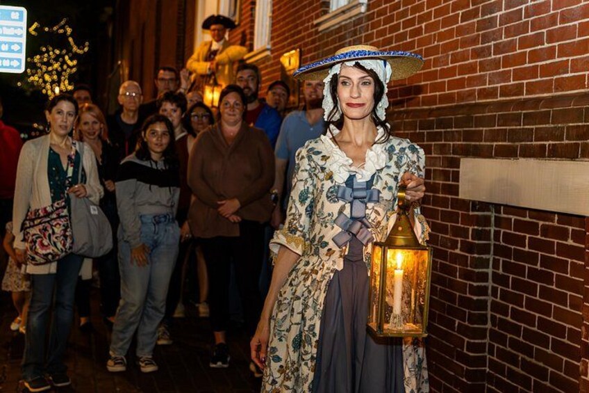 A person in historical attire holding a lantern, standing in front of a brick building with a crowd in the background. 1
