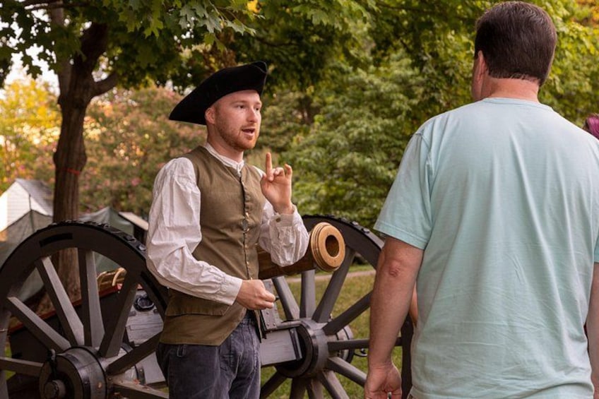 A person in historical costume standing next to a cannon, with trees and a tent in the background. 3