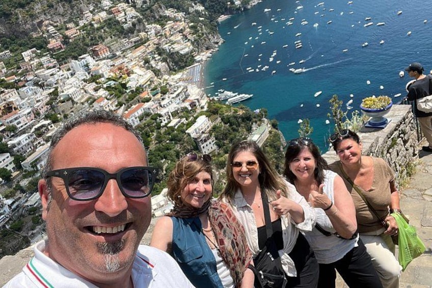 En grupp människor står på en klippa med utsikt över en kuststad, och nedanför dem ligger båtar i vattnet. 5