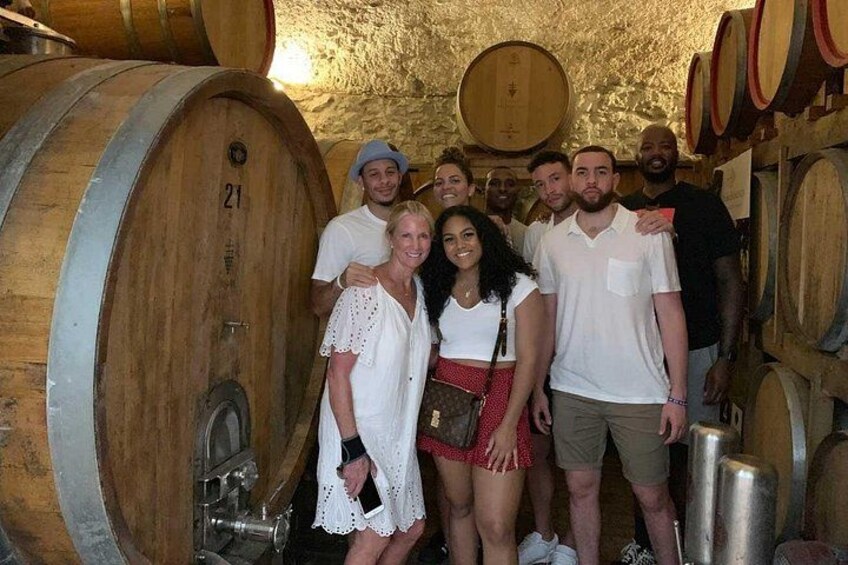 A group of people in a wine cellar with wooden barrels and a metal barrel. 1