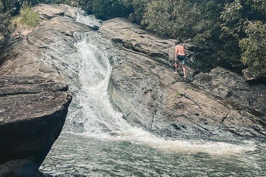 Ein Wasserfall stürzt über Felsen, eine Person steht auf einem Felsen, und im Hintergrund dichtes Grün. 5