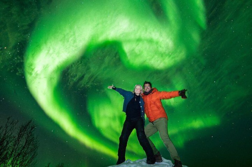 Two people posing with arms outstretched under a green aurora display. 1