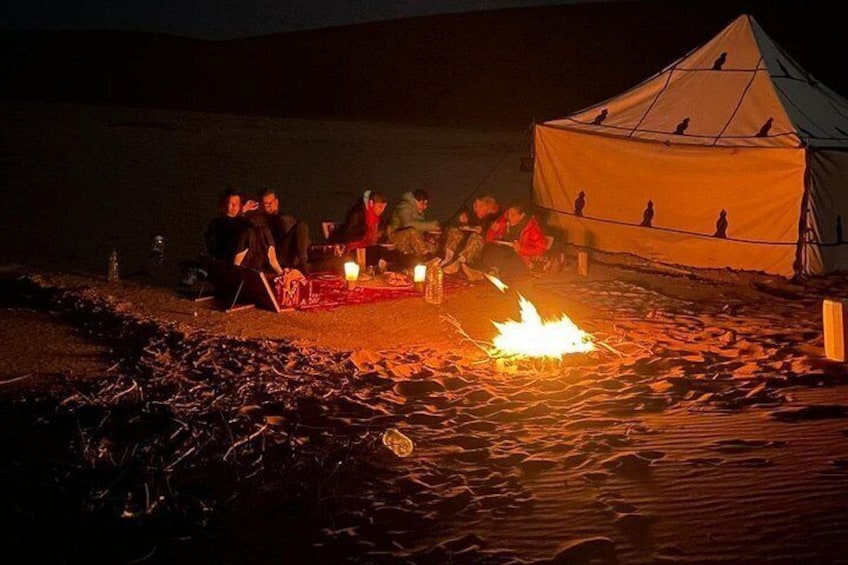Un groupe de personnes est assis autour d’un feu de camp, la nuit, avec une tente en arrière-plan. 4