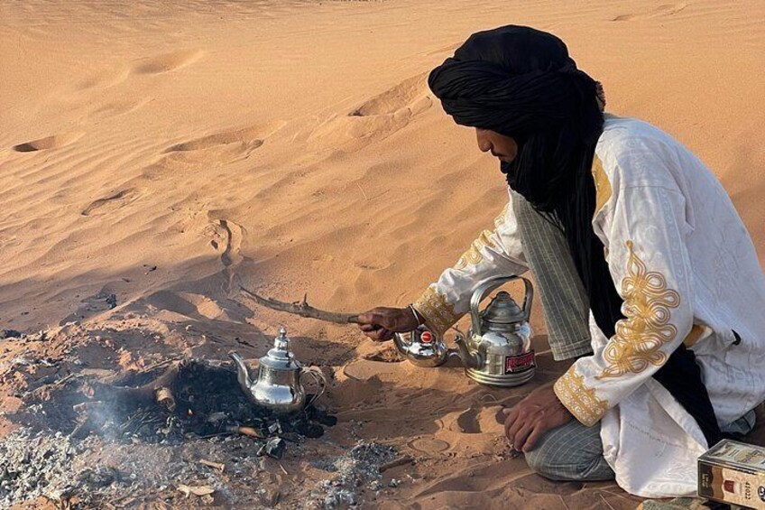 Une personne vêtue d’un costume traditionnel est assise sur le sable et prépare une boisson à l’aide d’une bouilloire en métal. 2