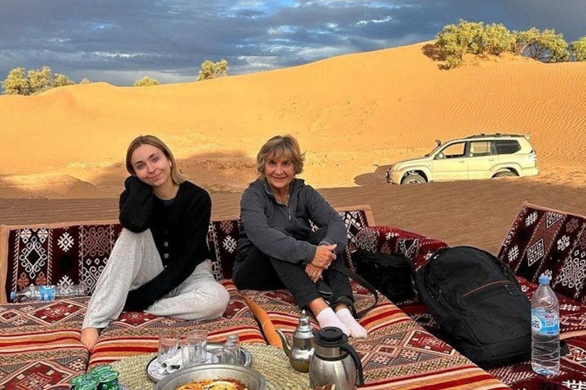 Dos personas sentadas sobre una alfombra estampada en un desierto con dunas de arena y un vehículo blanco al fondo. 1