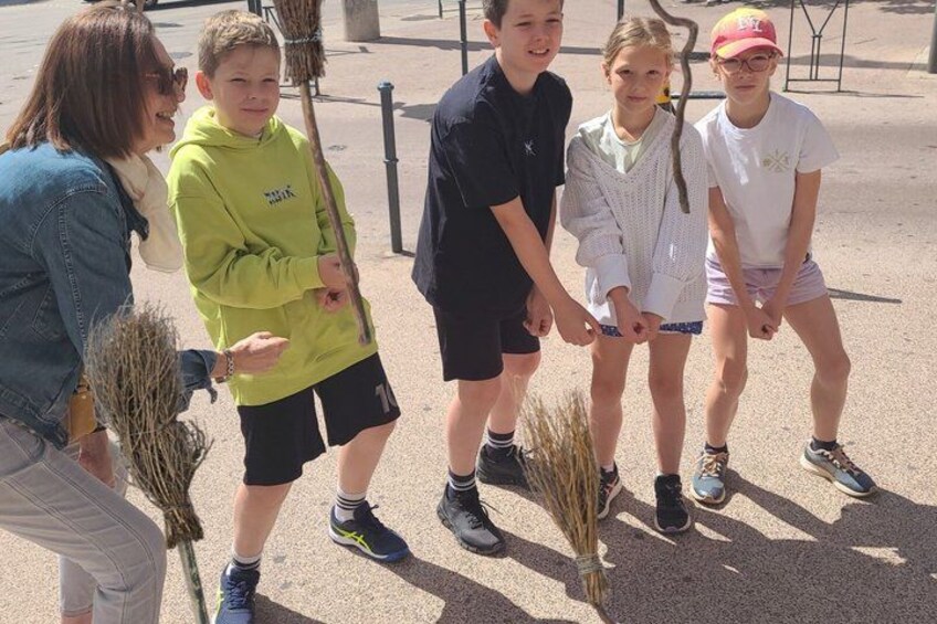 A group of children holding sticks and a broom, standing on a paved surface with a fence in the background. 2