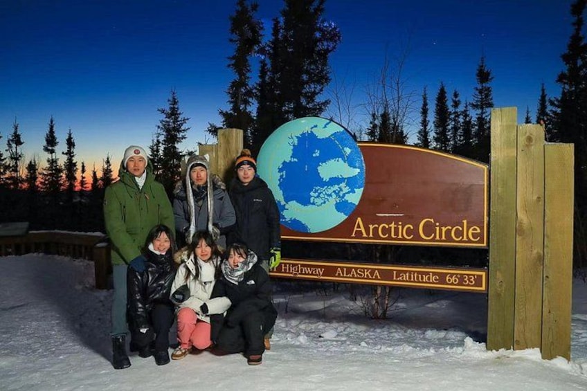 Una familia posando frente a un letrero que dice 'Círculo Ártico' con un gráfico de un globo terráqueo y referencias a la carretera Alaska Latitud 66°33. 4