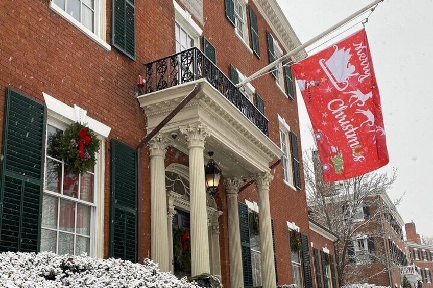 A brick building with a white porch and green shutters. A red banner with a Christmas tree and snowman is hanging on the right. 4