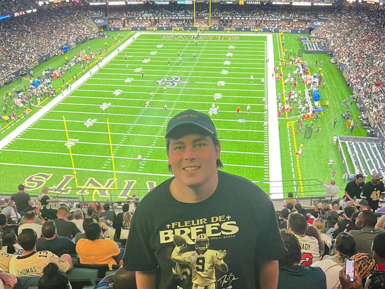 A football stadium with a green field marked with white lines, and a person wearing a black t-shirt with a football player graphic. 2