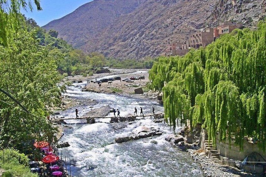 A river with people walking, a bridge, and buildings in the background. 1