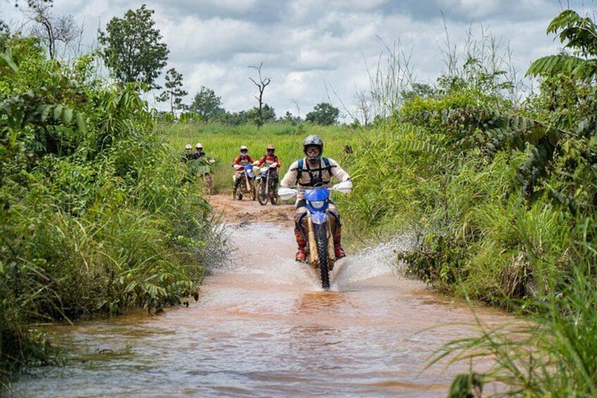 A group of motorcyclists riding through a shallow water crossing surrounded by greenery. 2