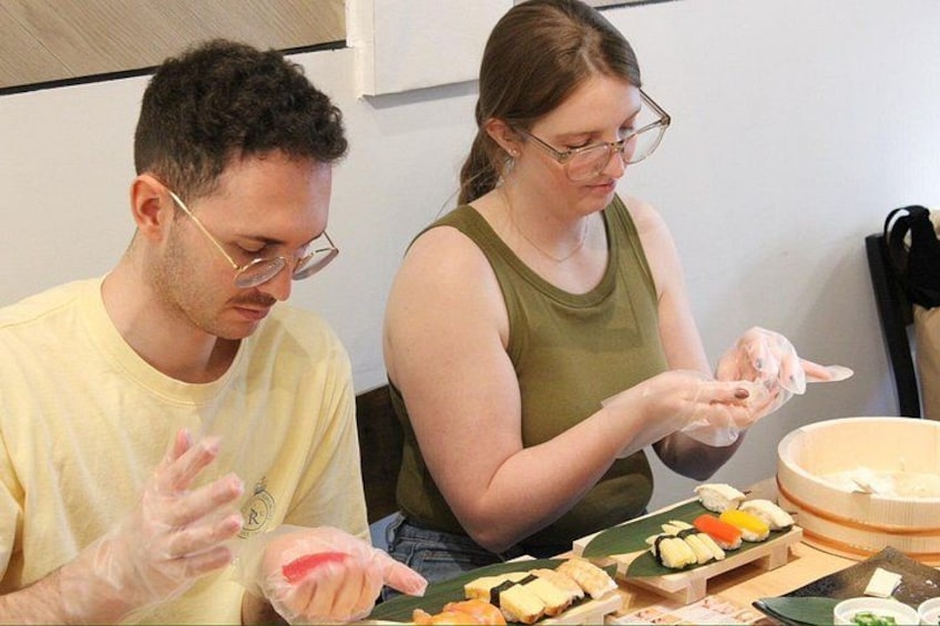 Dos personas preparando comida, probablemente sushi, con varios ingredientes y utensilios sobre la mesa. 2