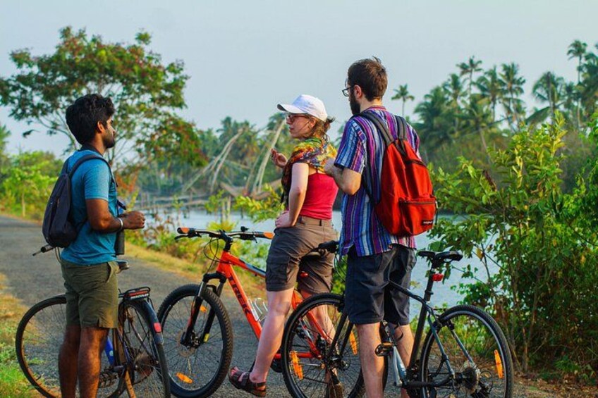 Three people standing by bicycles on a road with a water body and greenery in the background. 2