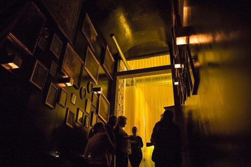 A dimly lit corridor with framed pictures on the walls and a group of people walking towards a bright area. 4