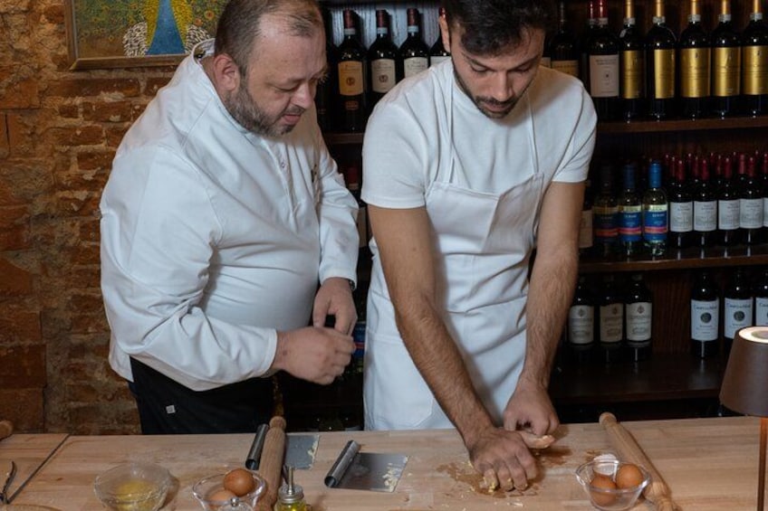 Two chefs preparing food in a kitchen with wine bottles in the background. 5