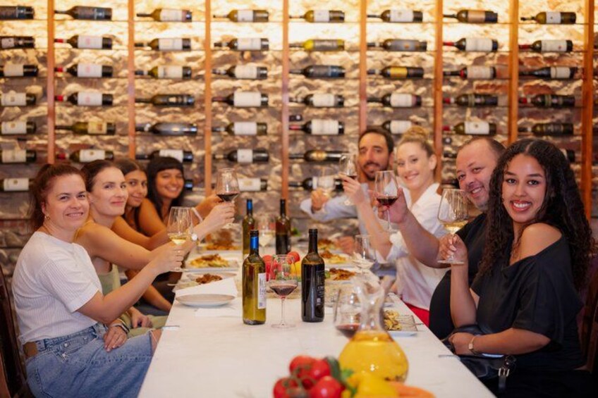 A group of people toasting with wine glasses at a table with a wine rack in the background. 3