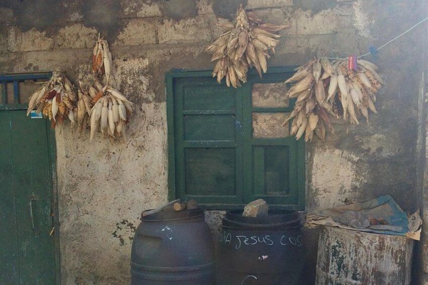 Un bâtiment rustique aux portes et aux murs verts, des bananes séchées suspendues au mur et deux grands tonneaux noirs portant l’inscription « JESUS CUS ». 4