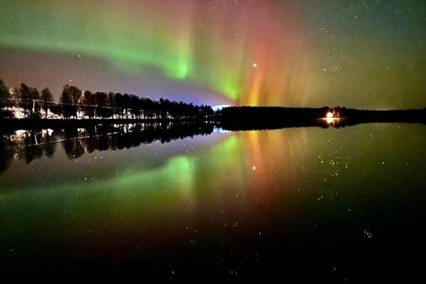 Een meer dat kleurrijke aurora's weerspiegelt, een oever met bomen, en een lichtbron in de verte. 2