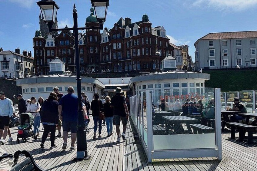 A wooden boardwalk with people walking, a glass enclosure with tables, and historic buildings in the background. 2