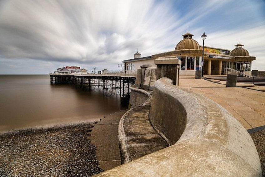 A coastal promenade with a pier extending into the water, a curved concrete barrier, and a building with domed structures. 5
