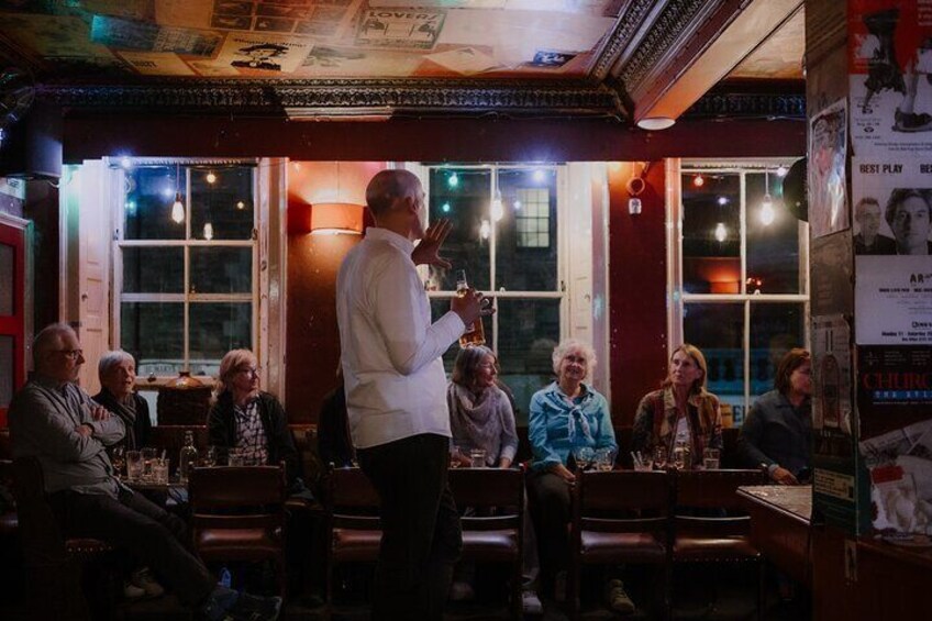 A person speaking to a group of seated individuals in a dimly lit room with wooden chairs and tables. 3
